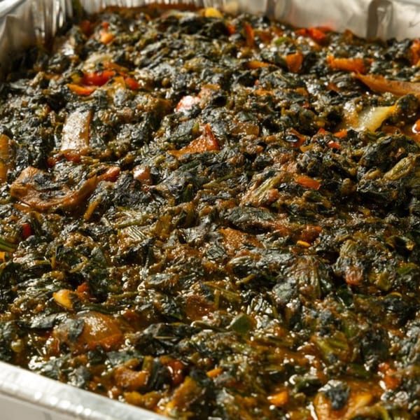 Close-up of a large aluminum tray filled with dark green African spinach stew, with hints of red and orange.