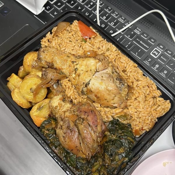 African jollof rice with chicken, plantains, and greens in a black takeout container next to a laptop.